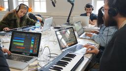 young people sat around a table with laptops and a keyboard making music 